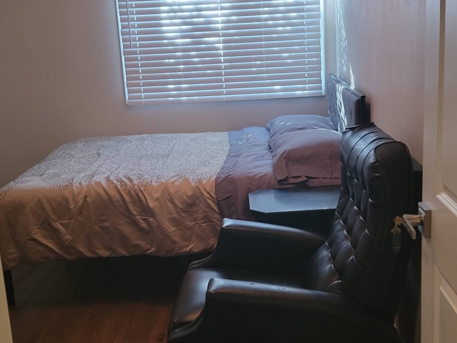 A resident bedroom with a made bed under the window, a side table and a leather armchair.