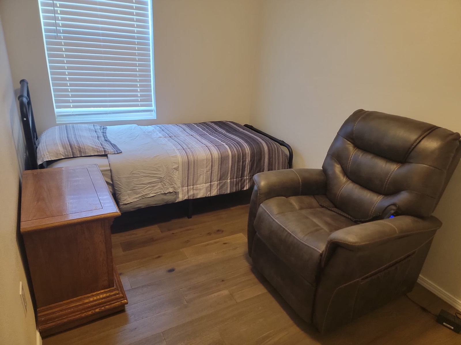 A resident bedroom with a made single bed under the window, a wooden nightstand and a recliner chair.