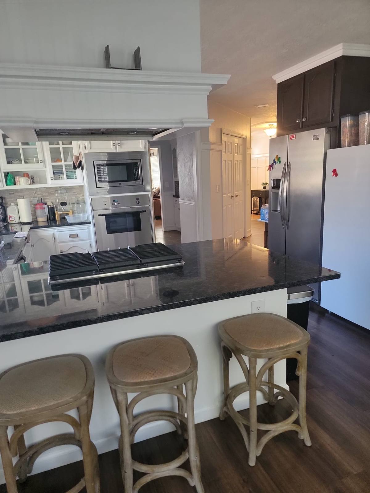 The kitchen counter with three bar stools, a wall oven and a stainless-steel fridge.