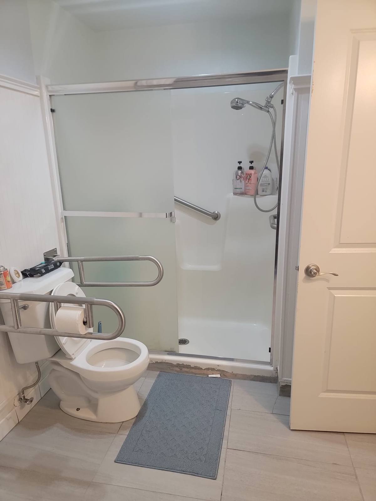 A shower with a handheld shower head and grab bar, next to a toilet with fold-down grab bars.